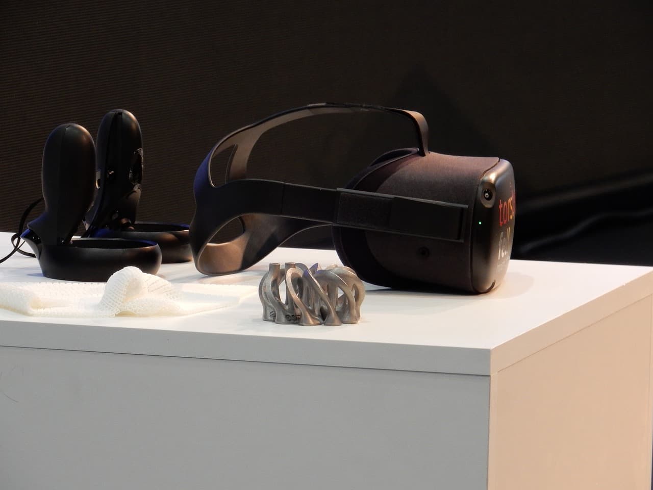 A virtual reality headset with two controllers sitting next to it all on top of a while table inside a dimly lit black room.