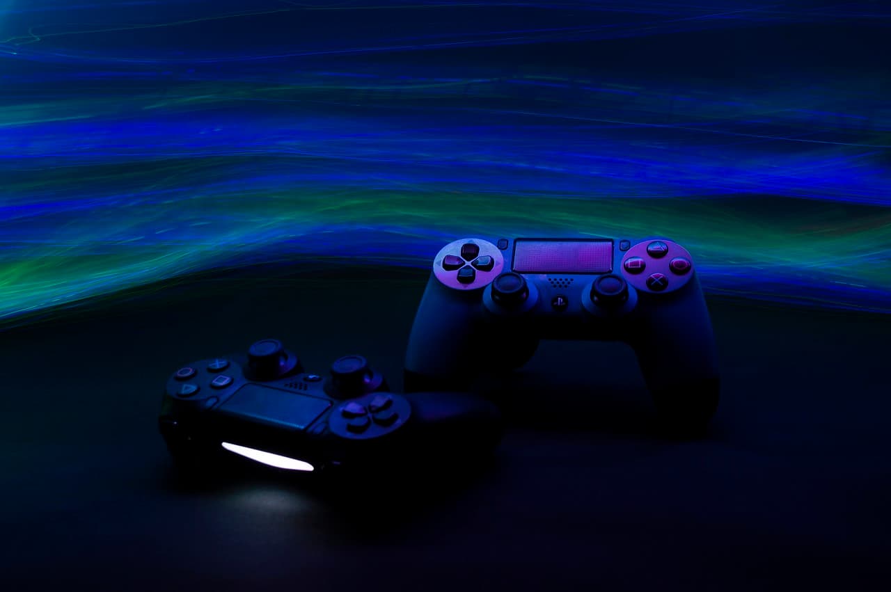 Two video game controllers on a black table in a dark room with a green and blue flow of lines that are lit up passing by them.