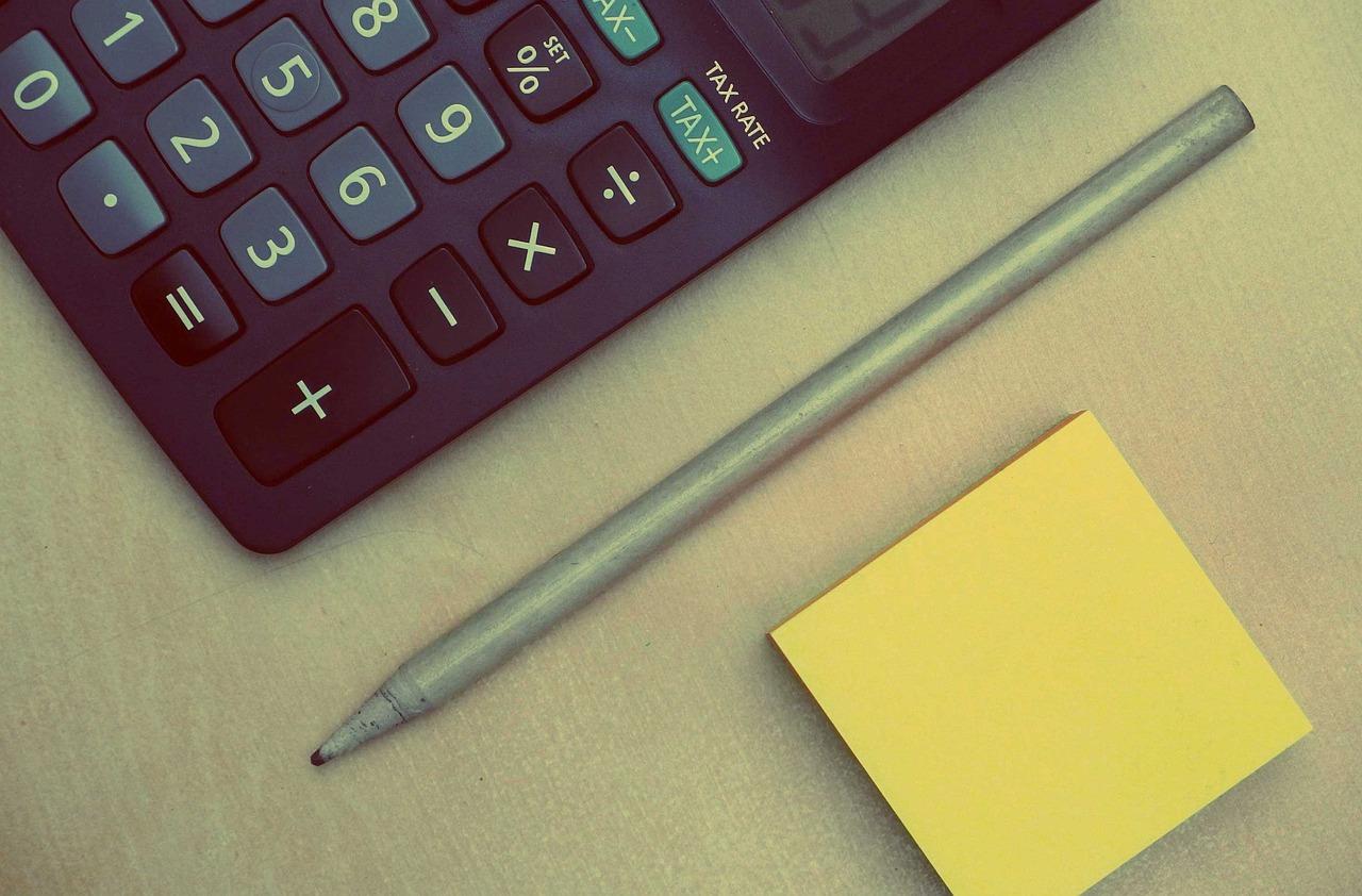 A gray pen without a cap next to a black calculator with white lettered buttons and a yellow sticky note pad all on a table.
