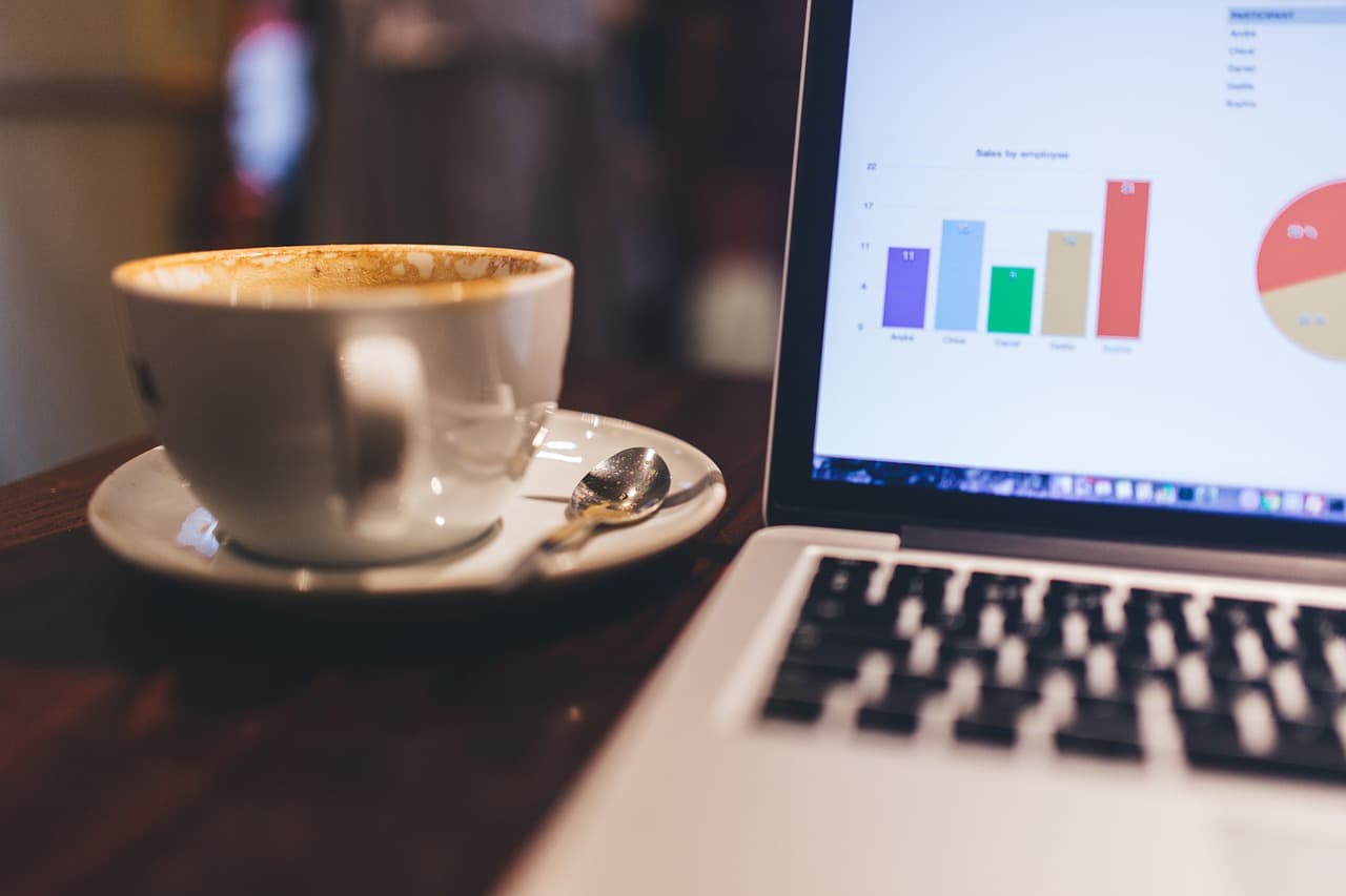 A white cup of coffee on a white place is sitting next to a gray macbook laptop with multi-colored bar and pie graphs on it.