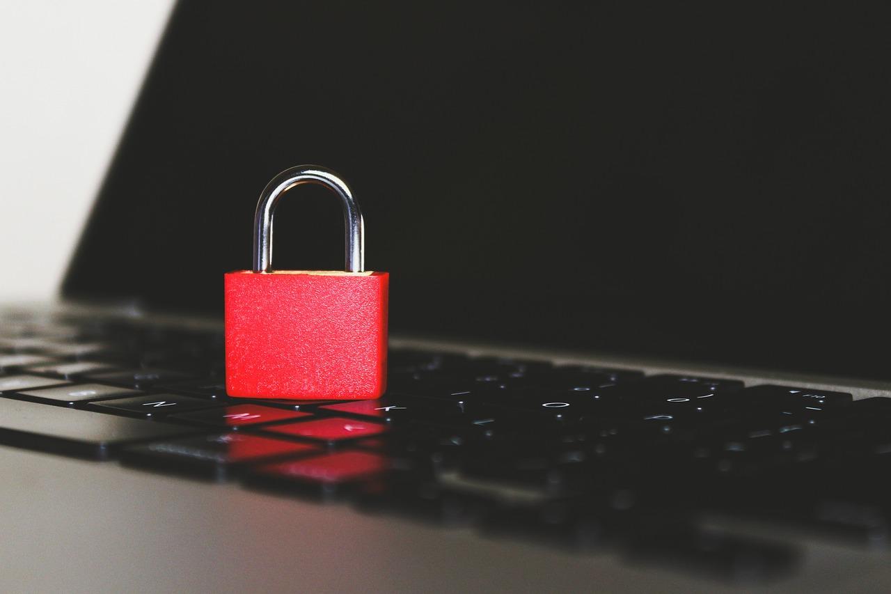A red padlock sitting on the keyboard of a dark gray macbook laptop that has a turned off black screen in front of a white wall.