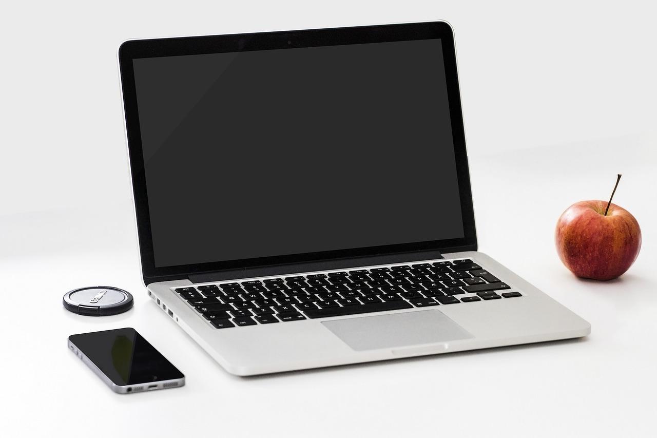 A gray macbook laptop with black keys on a white table next to an silver iPhone that has a turned off screen and a red apple.