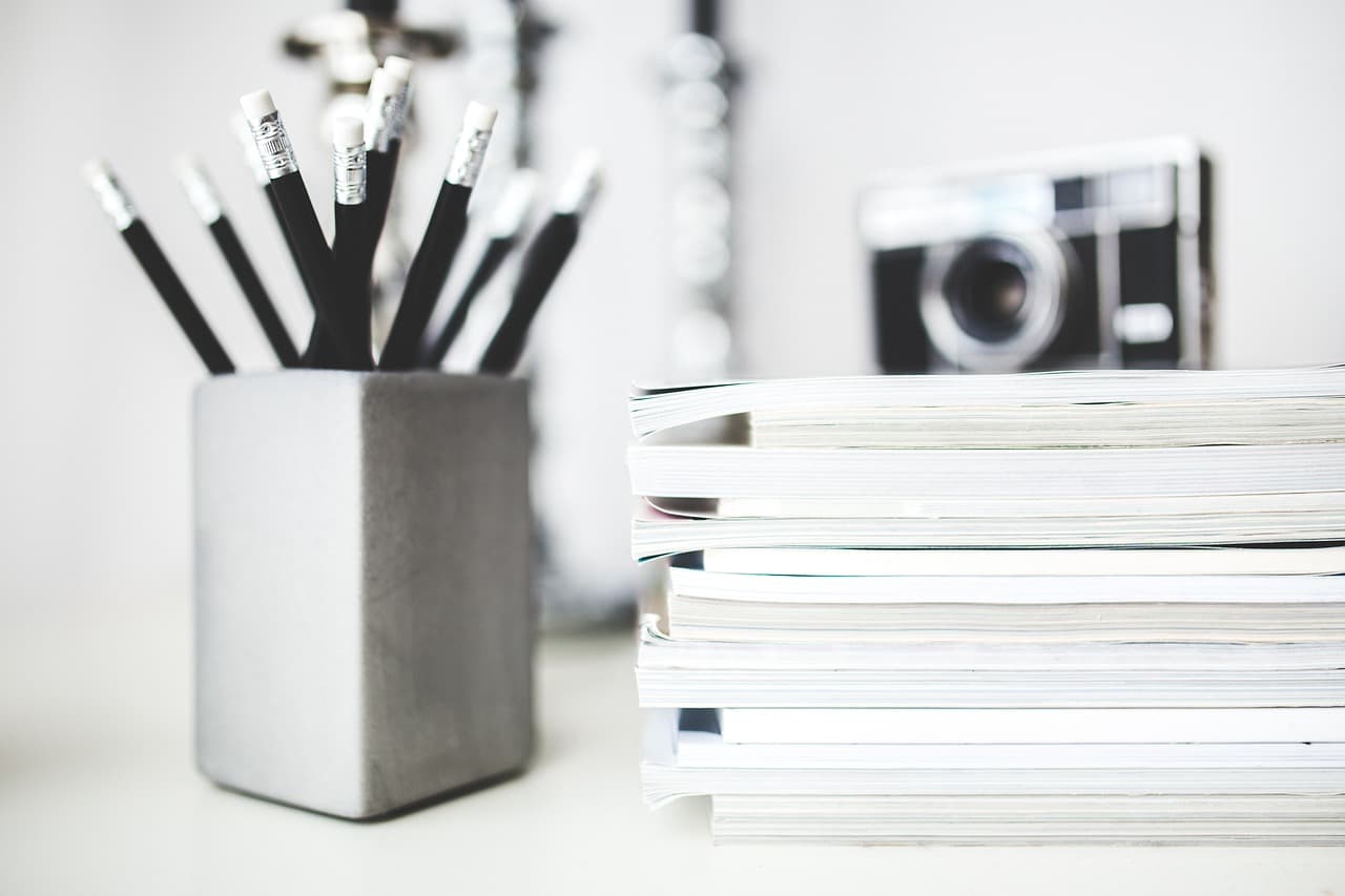 A silver matte colored tin that is holding multiple black pencils with white erasers on a white table next to a pile of books.