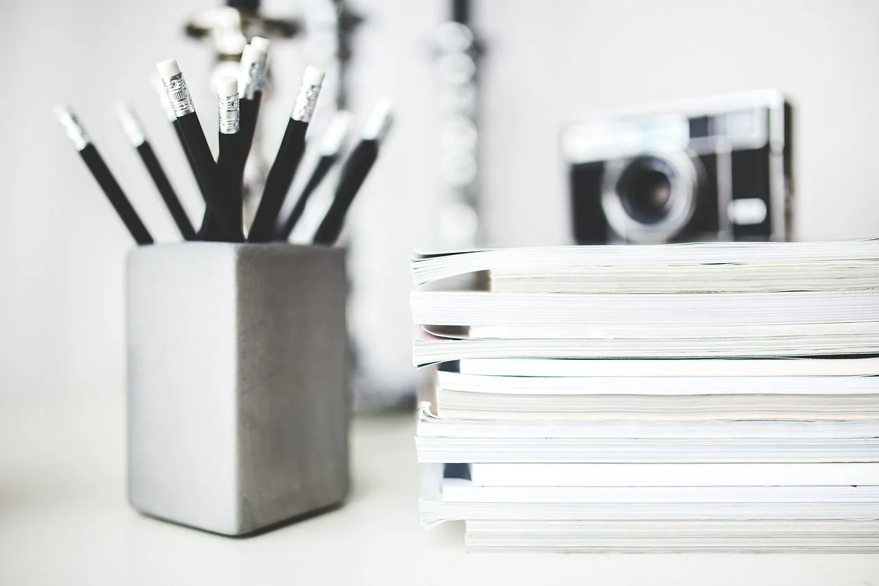 A silver matte colored tin that is holding multiple black pencils with white erasers on a white table next to a pile of books.
