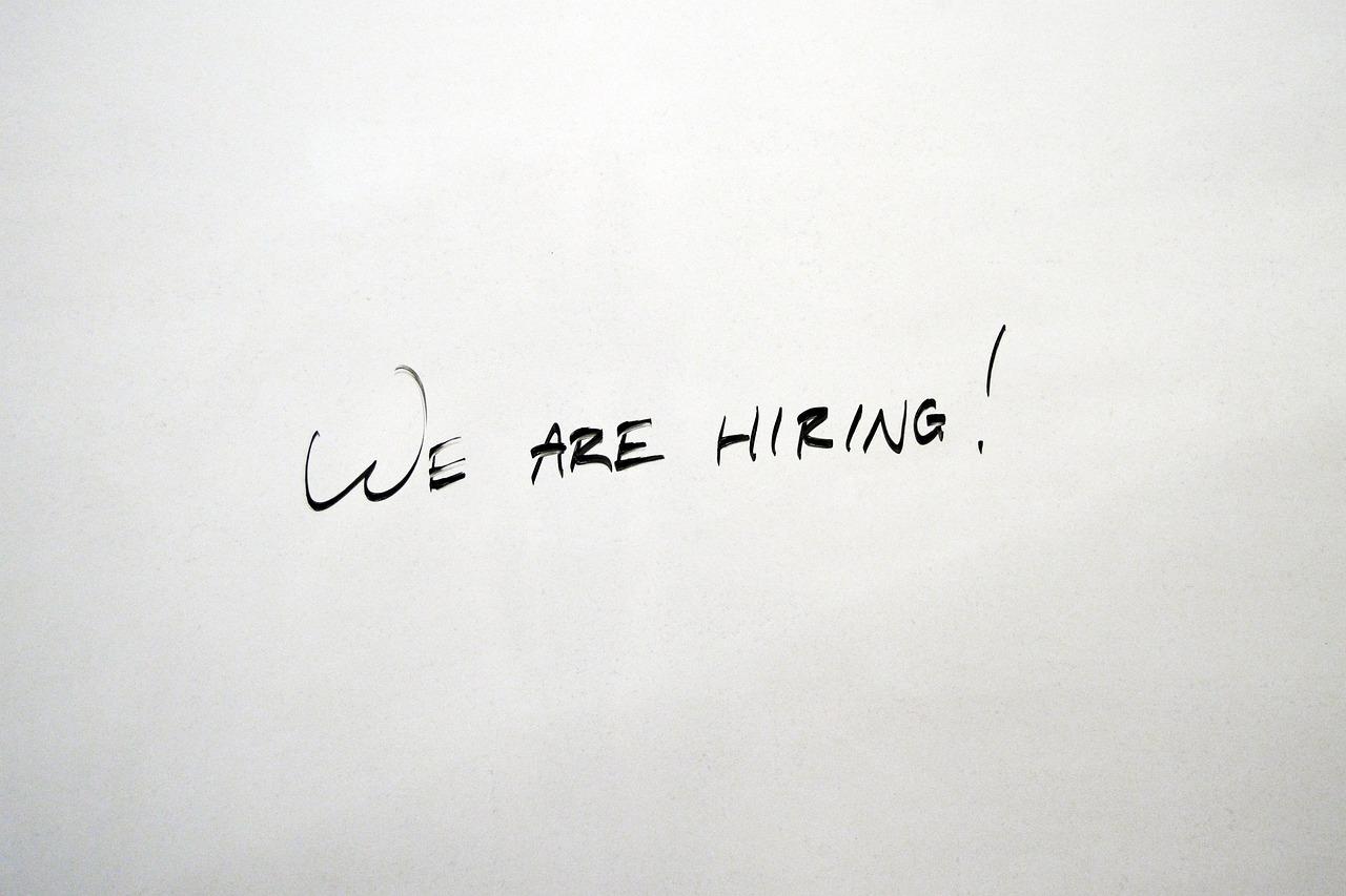 A white piece of paper that has written on it we are hiring with an exclamation mark all in black marker that is slightly faded.