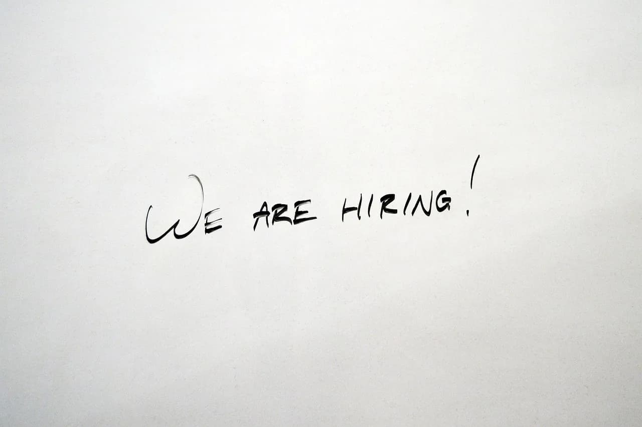 A white piece of paper that has written on it we are hiring with an exclamation mark all in black marker that is slightly faded.