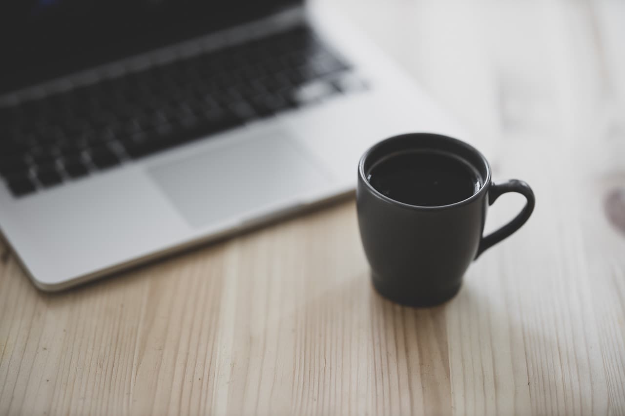 A dark green mug with black coffee in it on a lightly shaded wooden table that has a gray macbook laptop sitting next to it.