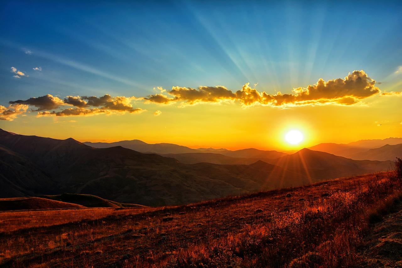 A mountain skyline with a sunset in the back of it with shining orange rays coming from it coloring the ground orange and red.