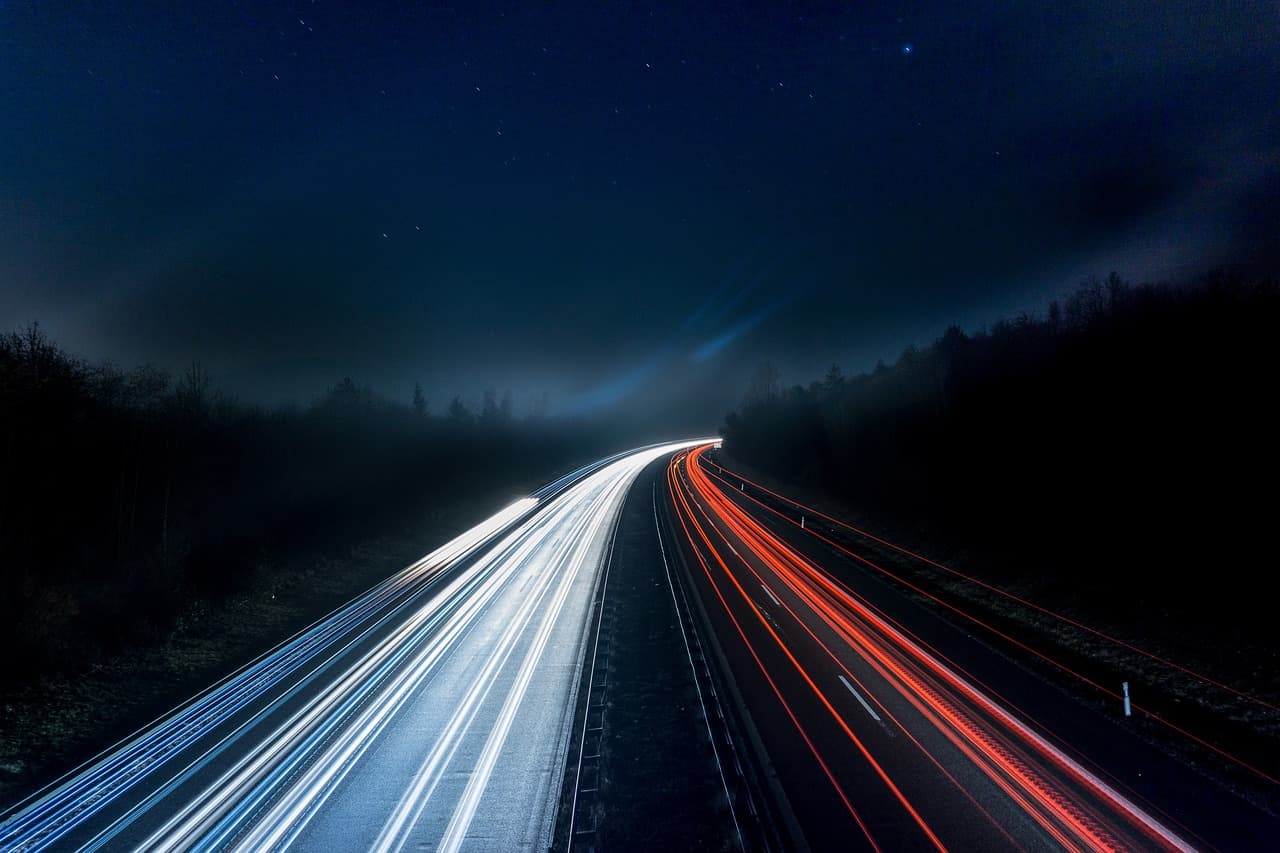 A dark highway with white stream headlights coming on the left side and red stream taillights going away on the right side.