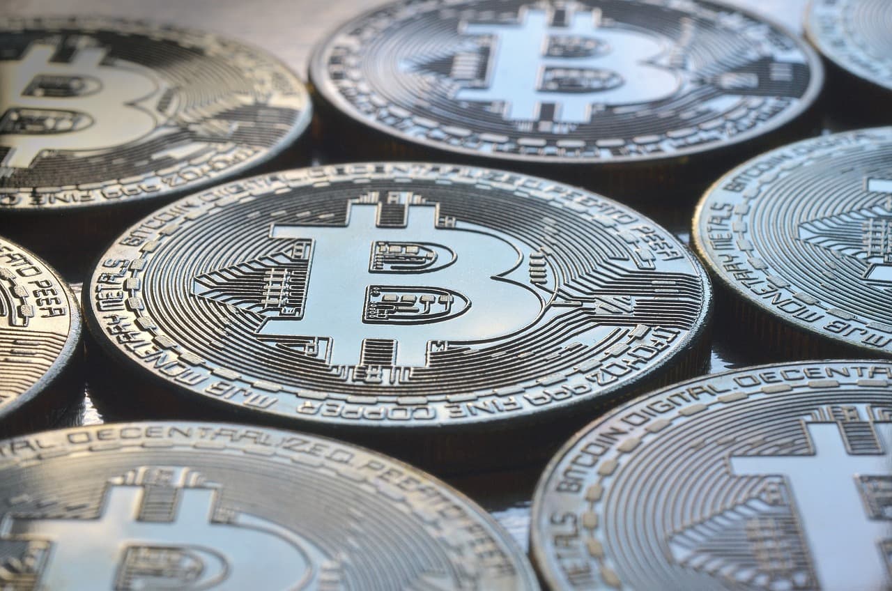 Eight golden coins on a table facing upwards, all with the Bitcoin logo engraved into the tops of them on a tan wooden table.