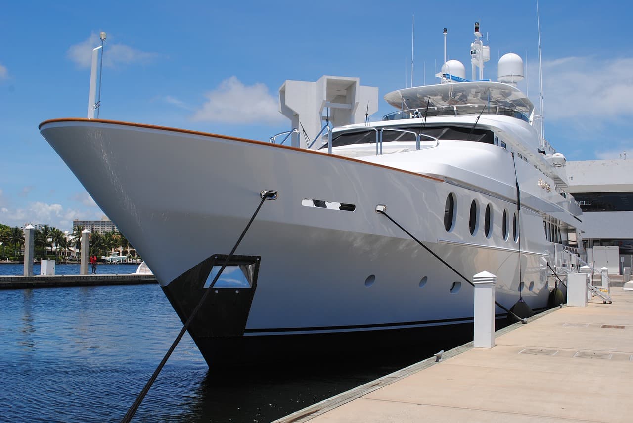 A giant white yacht on the blue ocean next to a tan cement-colored dock with a white bridge in the background of the ocean.