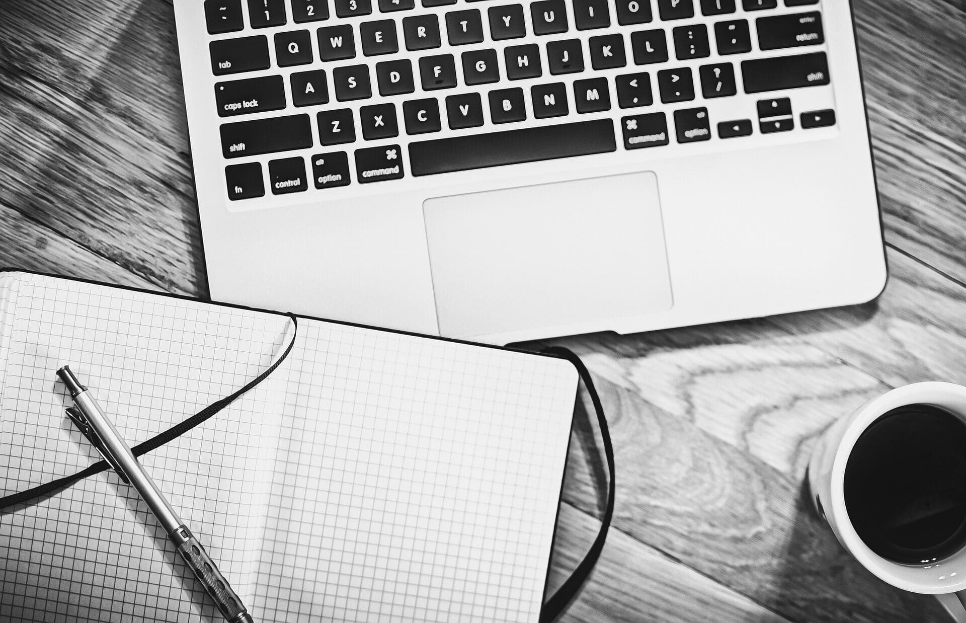 An Apple Macbook Laptop next to a notebook with a pen on it next to a cup of coffee on a wooden table all in black and white.