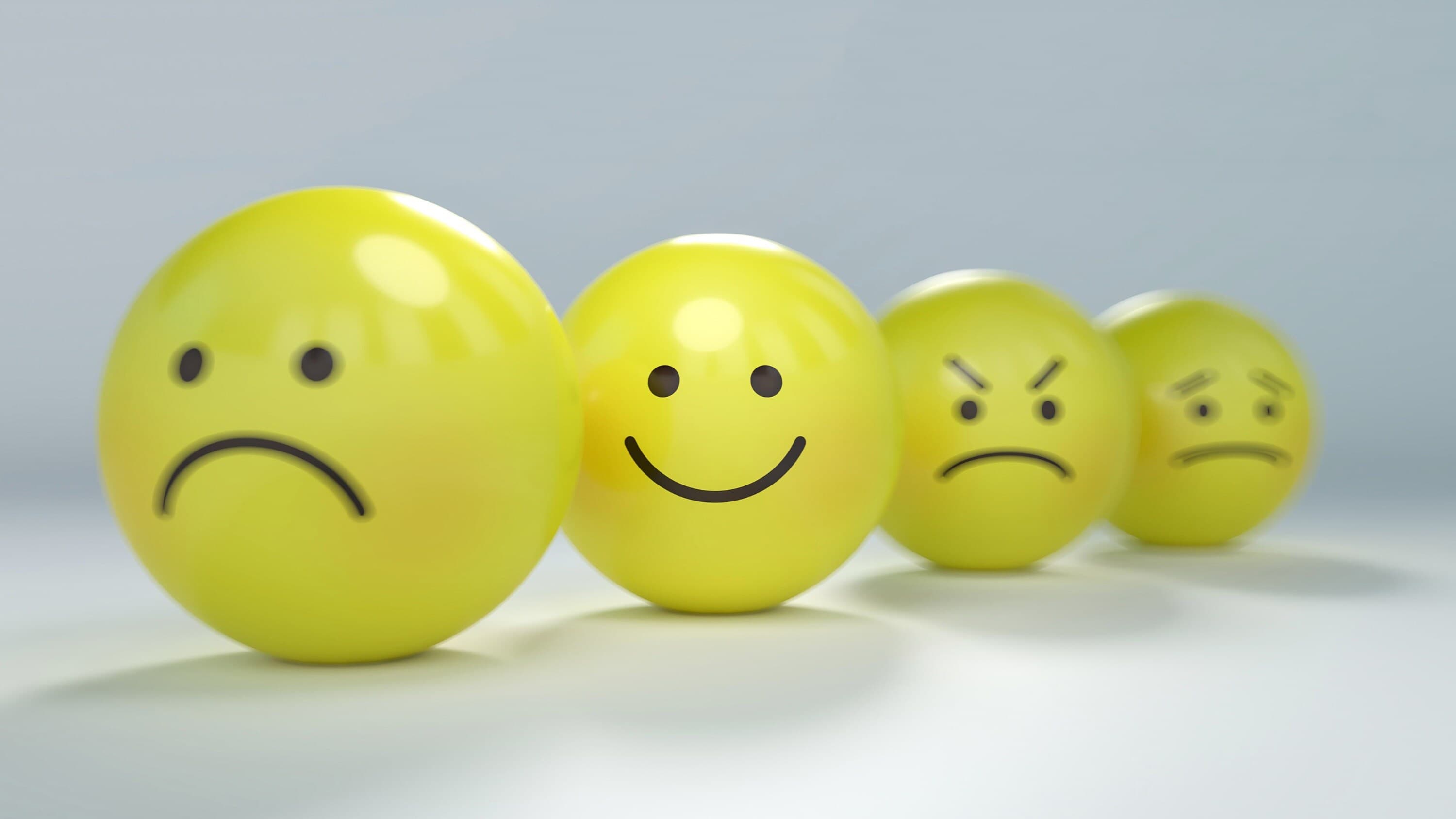 Many different yellow balls with faces on them showing different emotions with the focus on the happy one all on a white table.