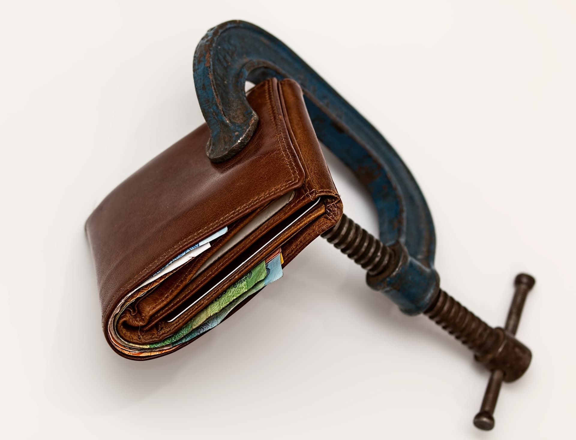 A leather wallet with cards and cash inside it being pinched together by a rustic green painted metal clamp on a white surface.