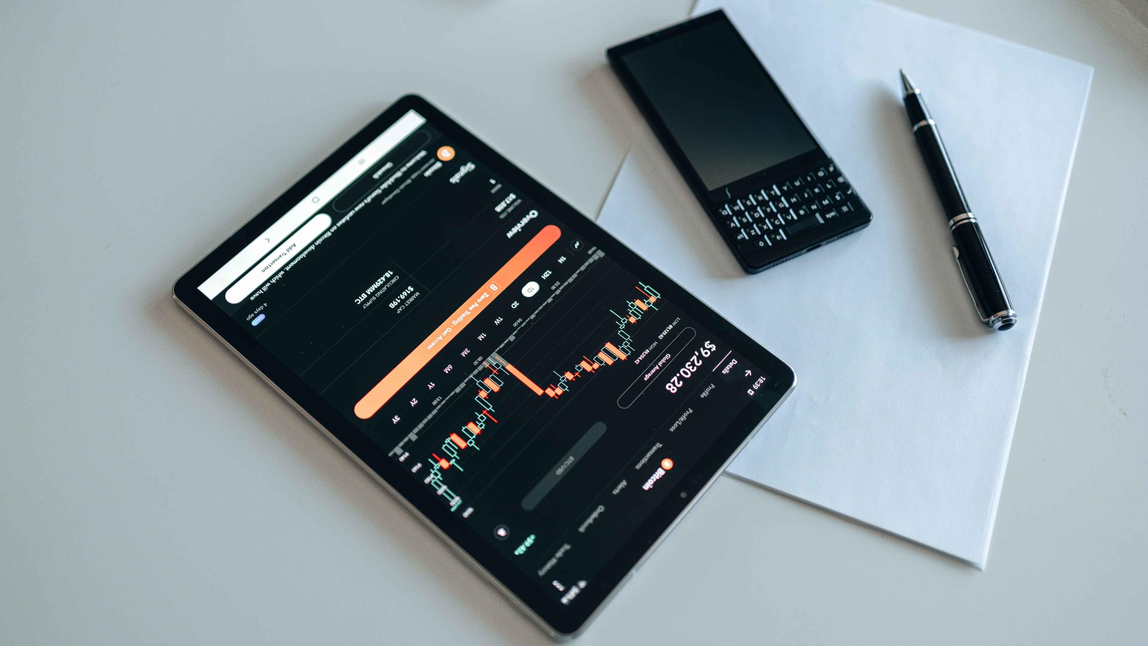 A black tablet with the crypto market on it next to a black phone and a black pen on a white piece of paper all on a table.