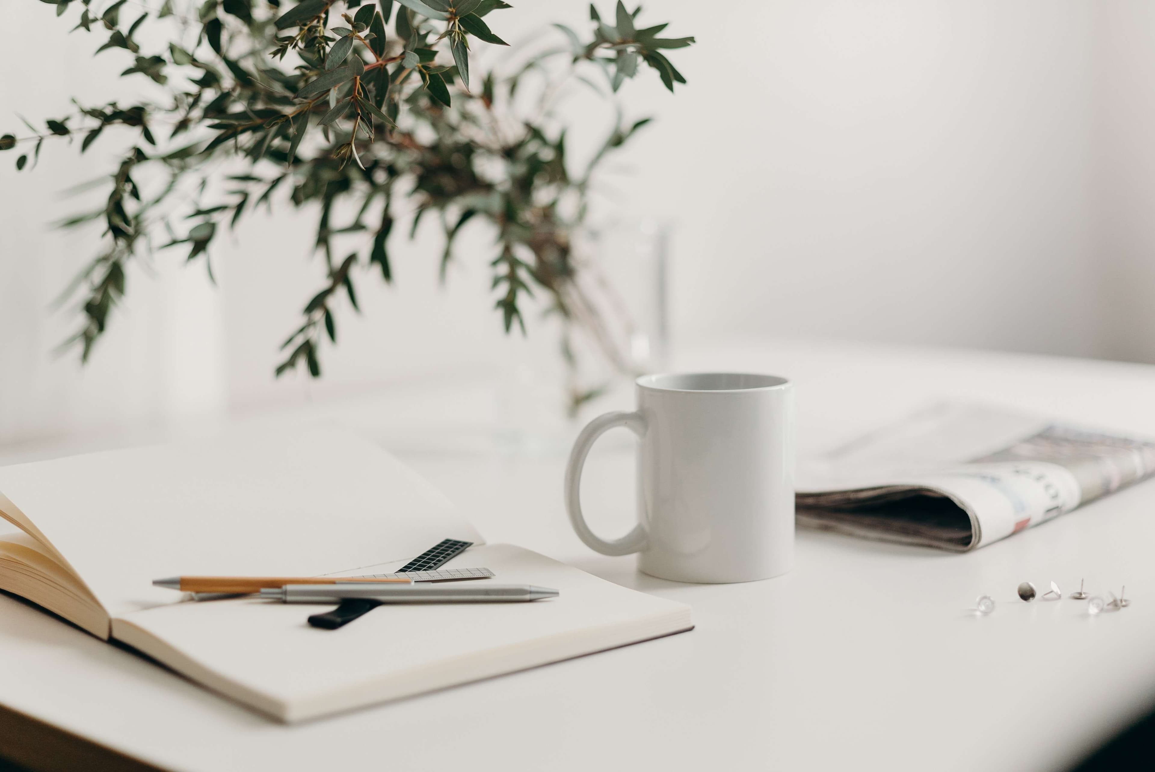 A newspaper on a white table next to a cup of coffee and a white lined paper notebook with rulers and pens resting on top of it.