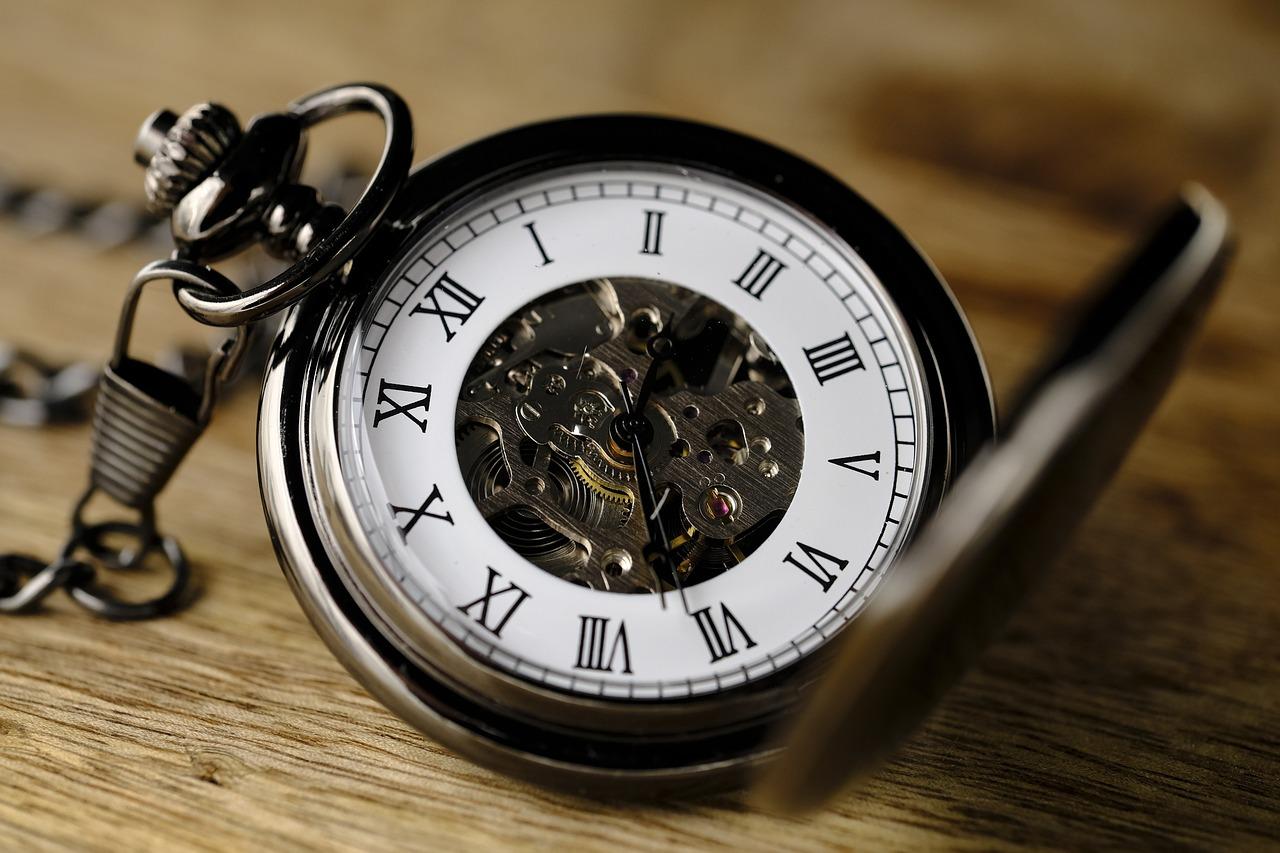 A pocket watch sitting on a wooden table with the gears exposed in the middle and a white background to the black hour numbers.