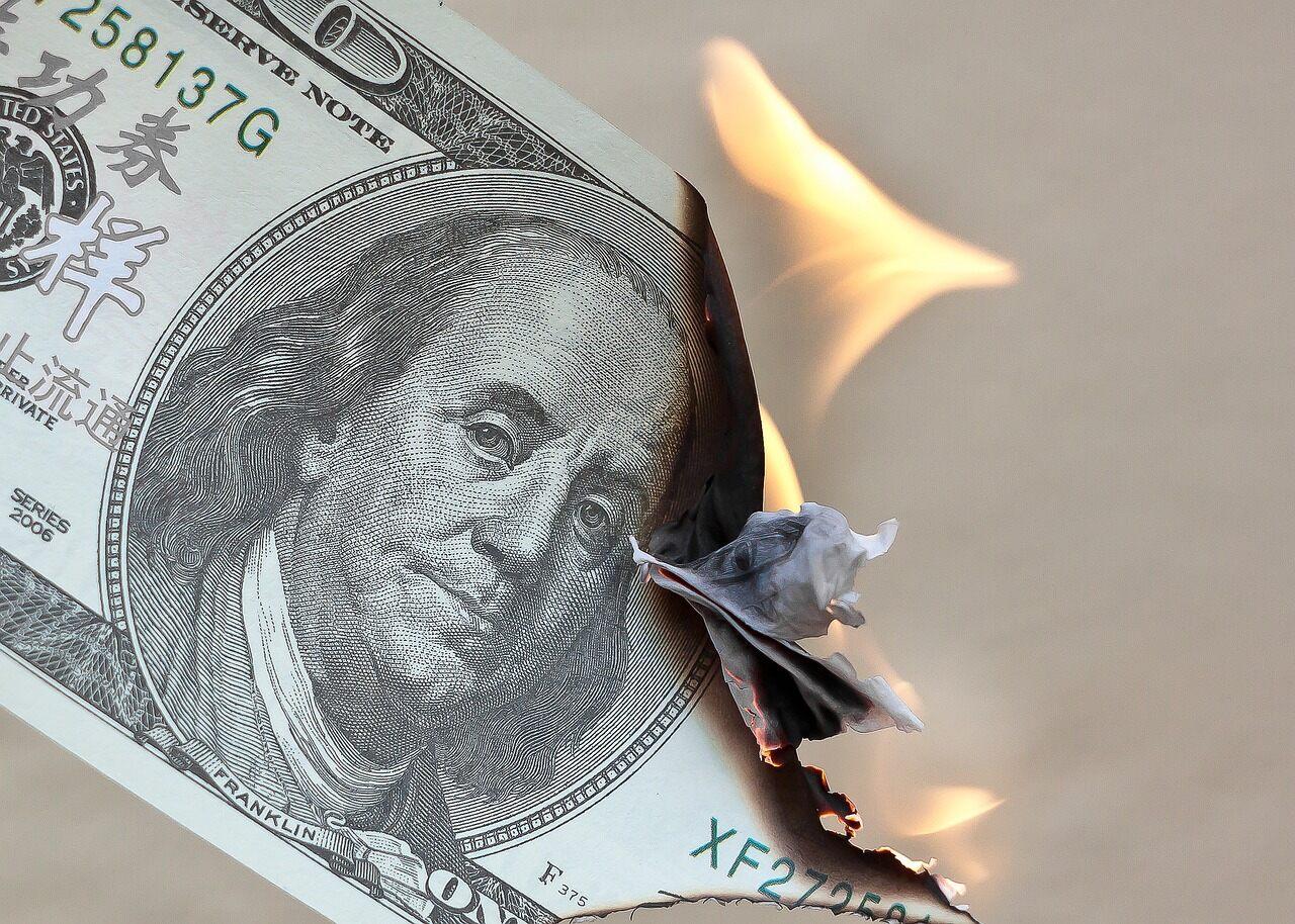 A one hundred dollar bill with the face side pointing forward on fire and almost halfway burnt up in front of a light surface.