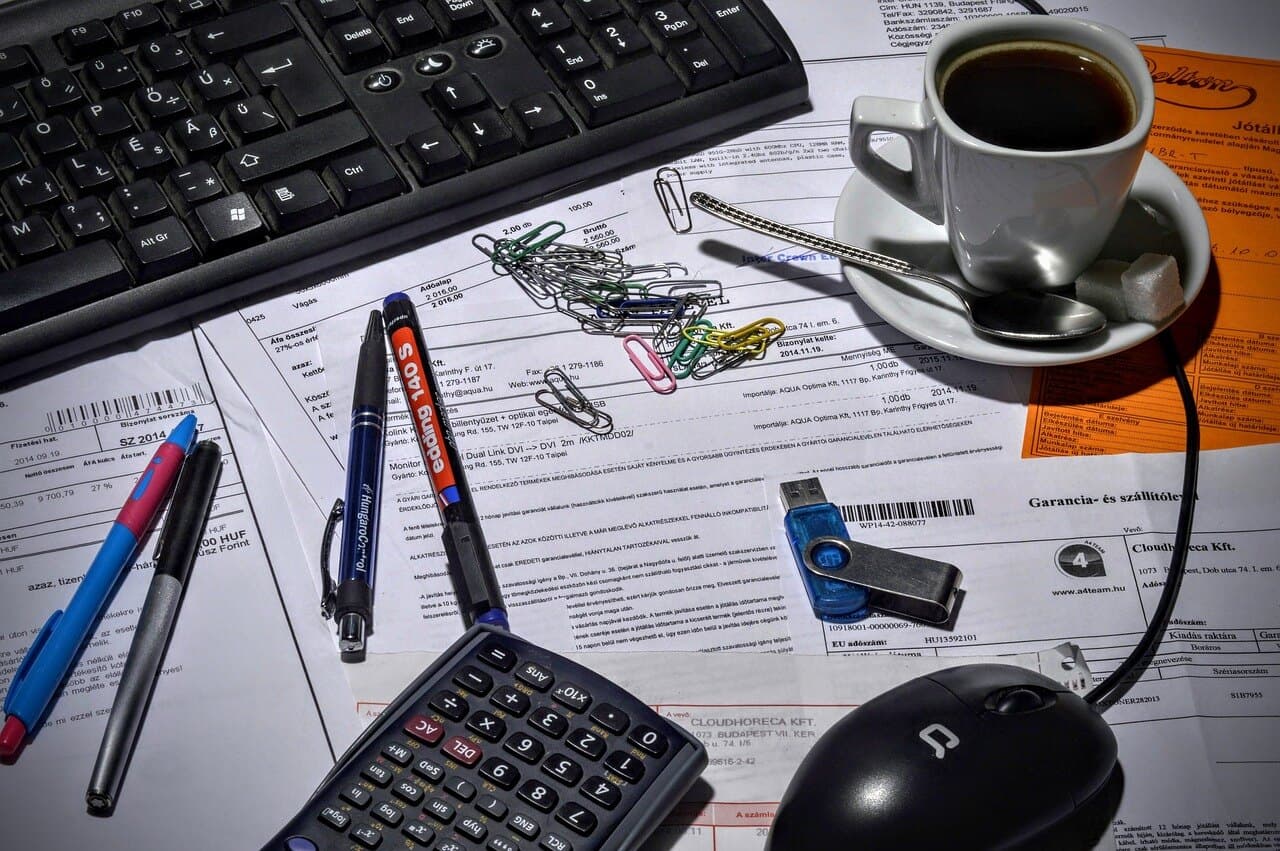 A pile of paper clips on a table with a bunch of papers covering it next to pens a cup of coffee, a calculator, and a mouse.