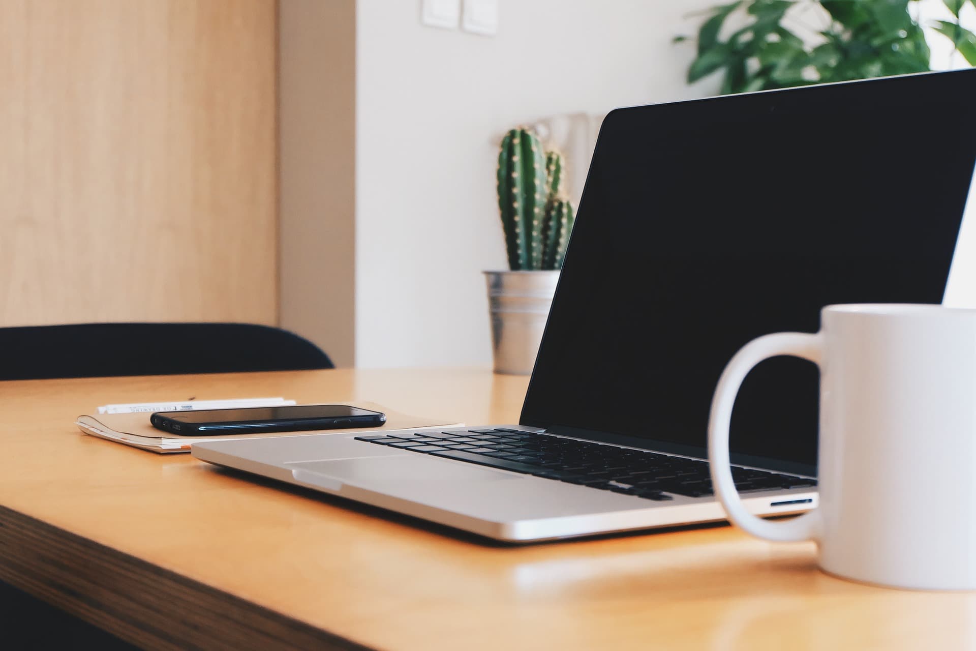 An Apple Macbook Laptop on a light wooden table next to a white coffee cup and a notebook with an Apple iPhone on top of it.