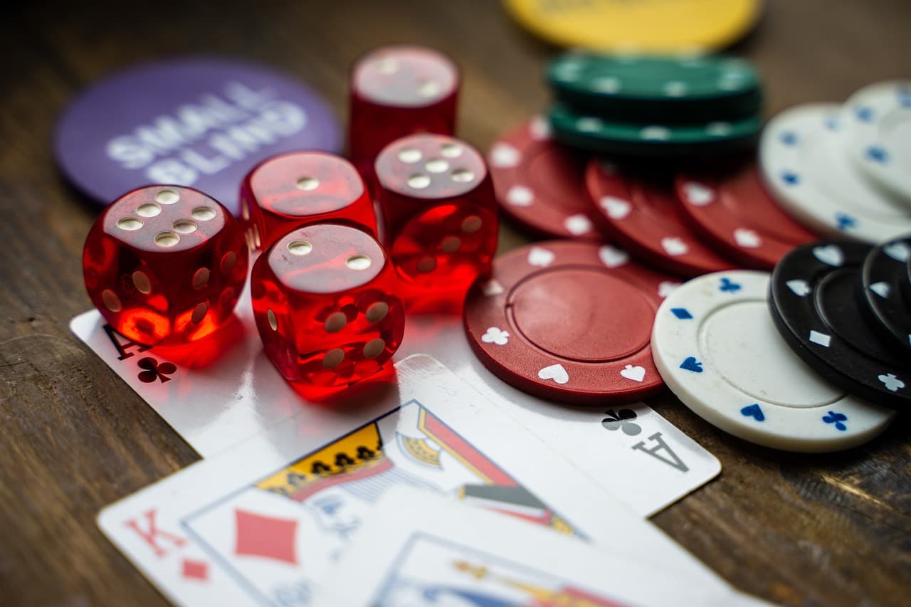 Five red see through dice with white dots resting on top of three playing cards next to a pile of multi colored poker chips.