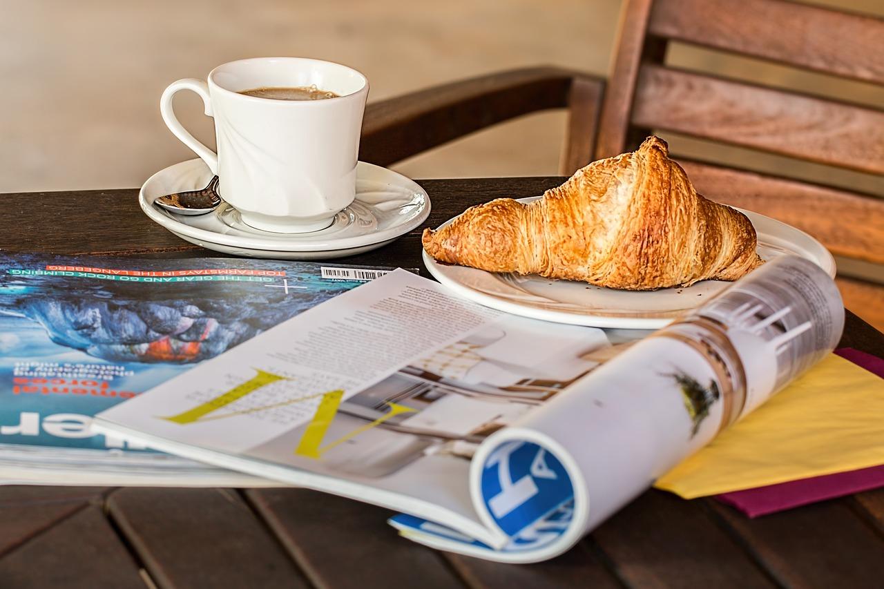 A coffee in a white mug and a croissant on a plate resting on a wooden diner table that has a rolled up newspaper on top of it.