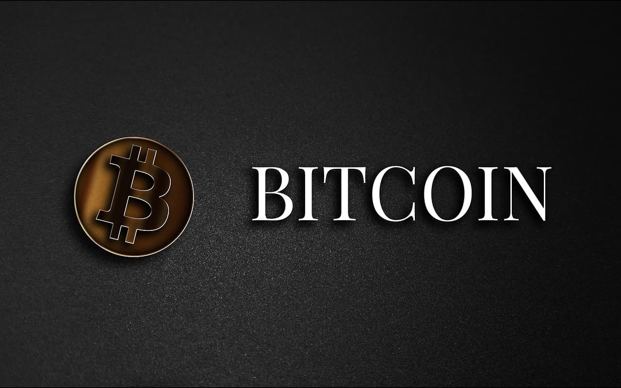 A golden coin with the Bitcoin logo on it in black next to the word Bitcoin all in front of a shadowy dark gray background.