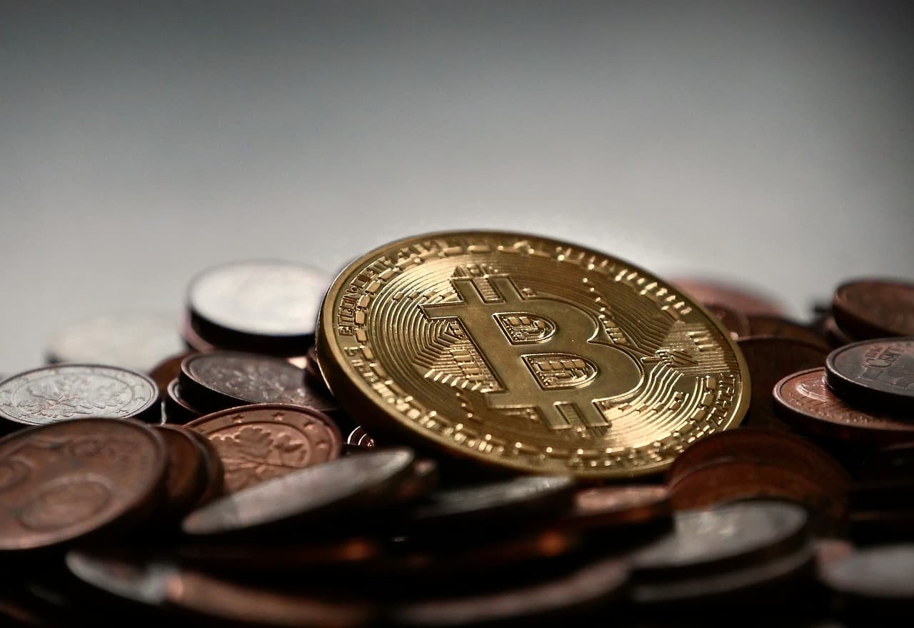 A golden coin on a stack of multiple other regular currency coins that has a bitcoin logo imprinted into it on a grayish table.