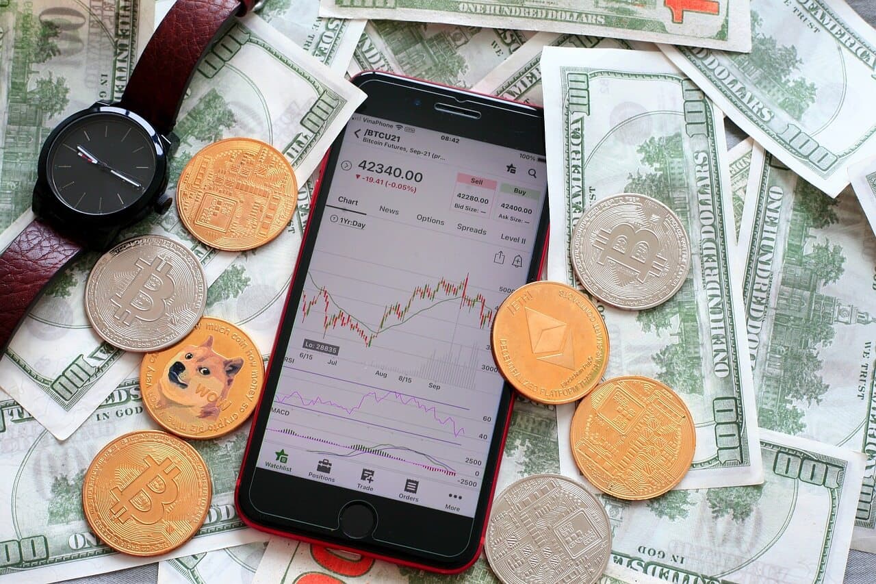 An Apple iPhone showing a crypto trading candle chart that is going up and down placed in top of a pile of money and coins.