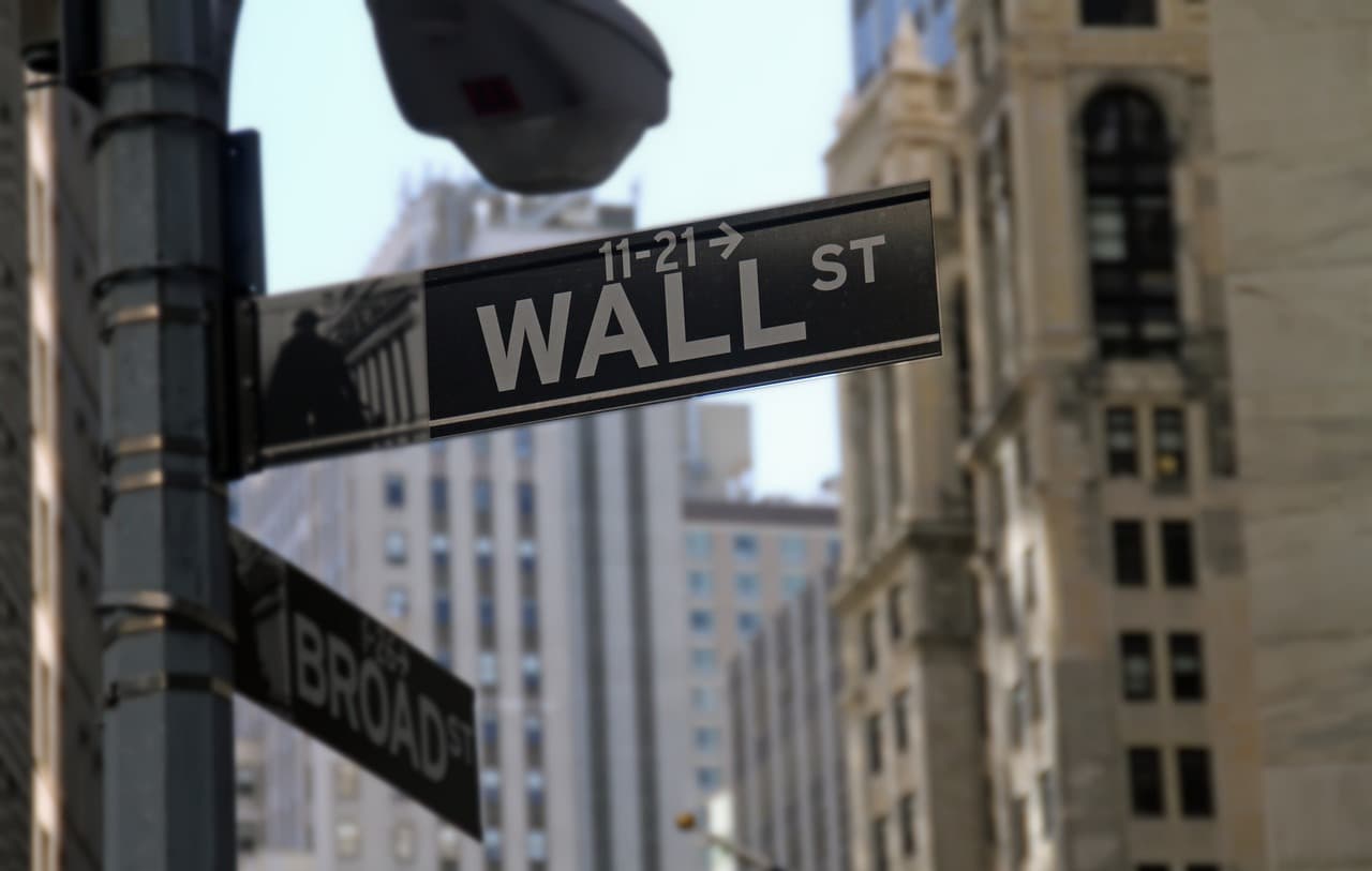 A black sign in a city labeled wall street in white letters with two large tan buildings in the background with black windows.