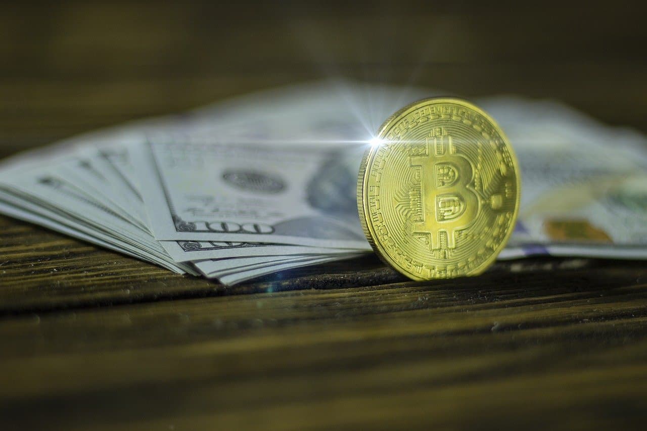 A stack of green hundred dollar bills on a brown wooden table with a golden coin showing the logo of Bitcoin on the front.