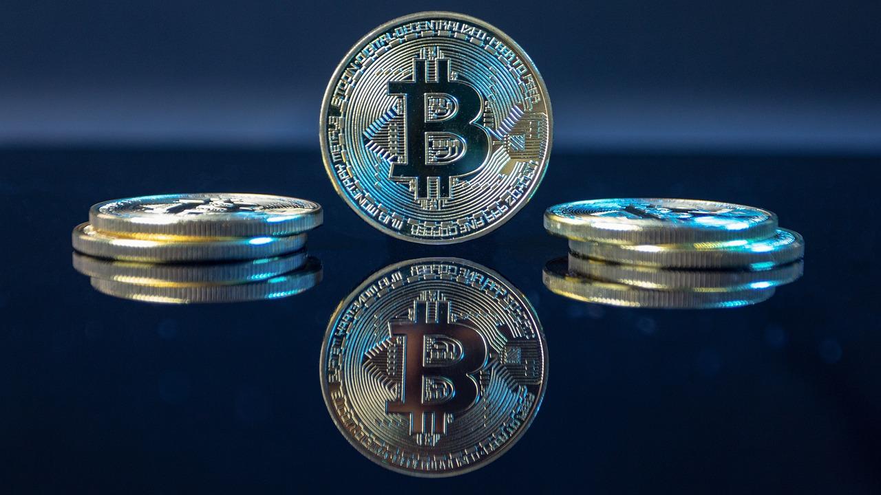 A golden coin with the Bitcoin logo is sitting up on a blue reflective table with its reflection below it with two other coins.