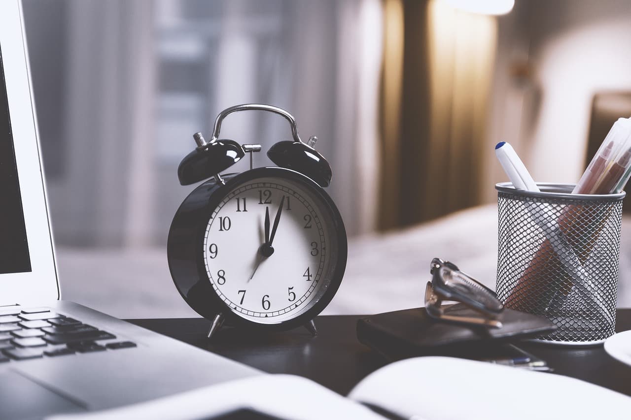 A black clock with a white face and black hands sitting on a light brown desk next to a grey laptop and white pencil holder.