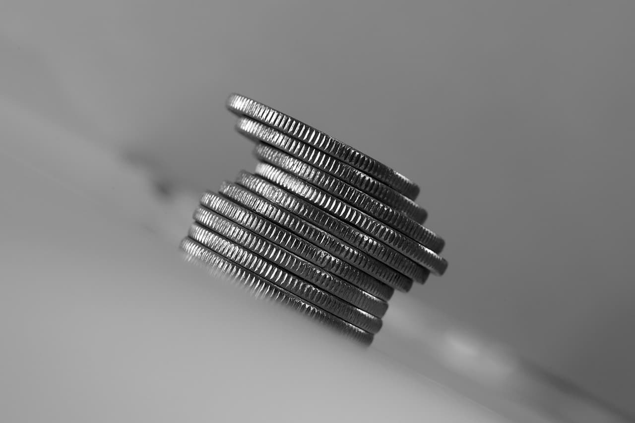 A stack of ten silver and shiny coins all on top of each other on a tilted white surface making the stack almost fall over.