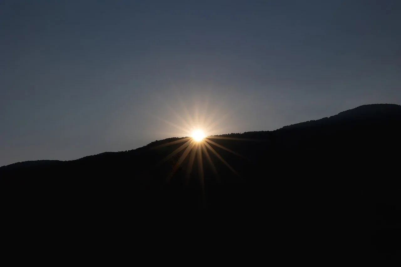A sunset that is right behind a dark black mountain shadow with the light yellow shining all over and brightening up the sky.