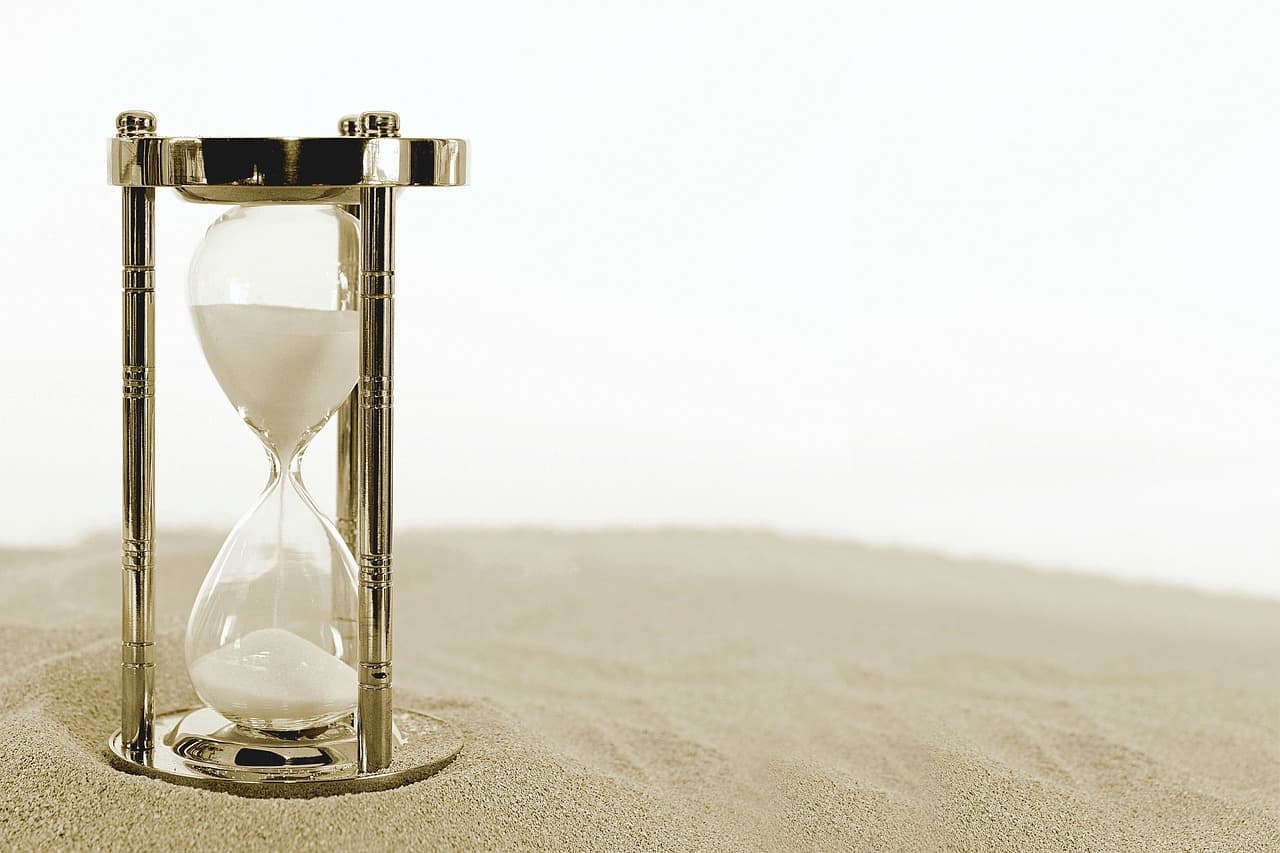 An hourglass with white sand inside it with golden support on the top and bottom along with golden beams on top of yellow sand.