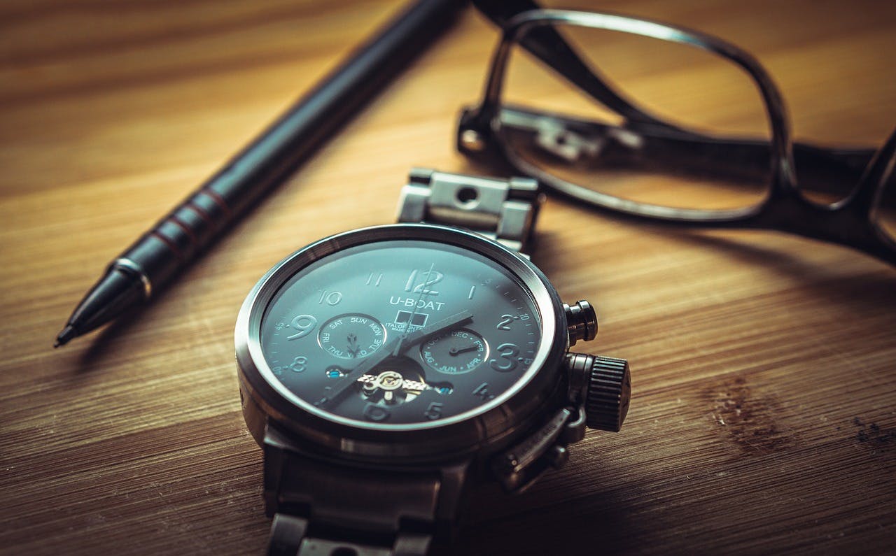 A black watch on a dark brown wooden table next to a black pen and a folded-up set of dark brown glasses behind the watch.