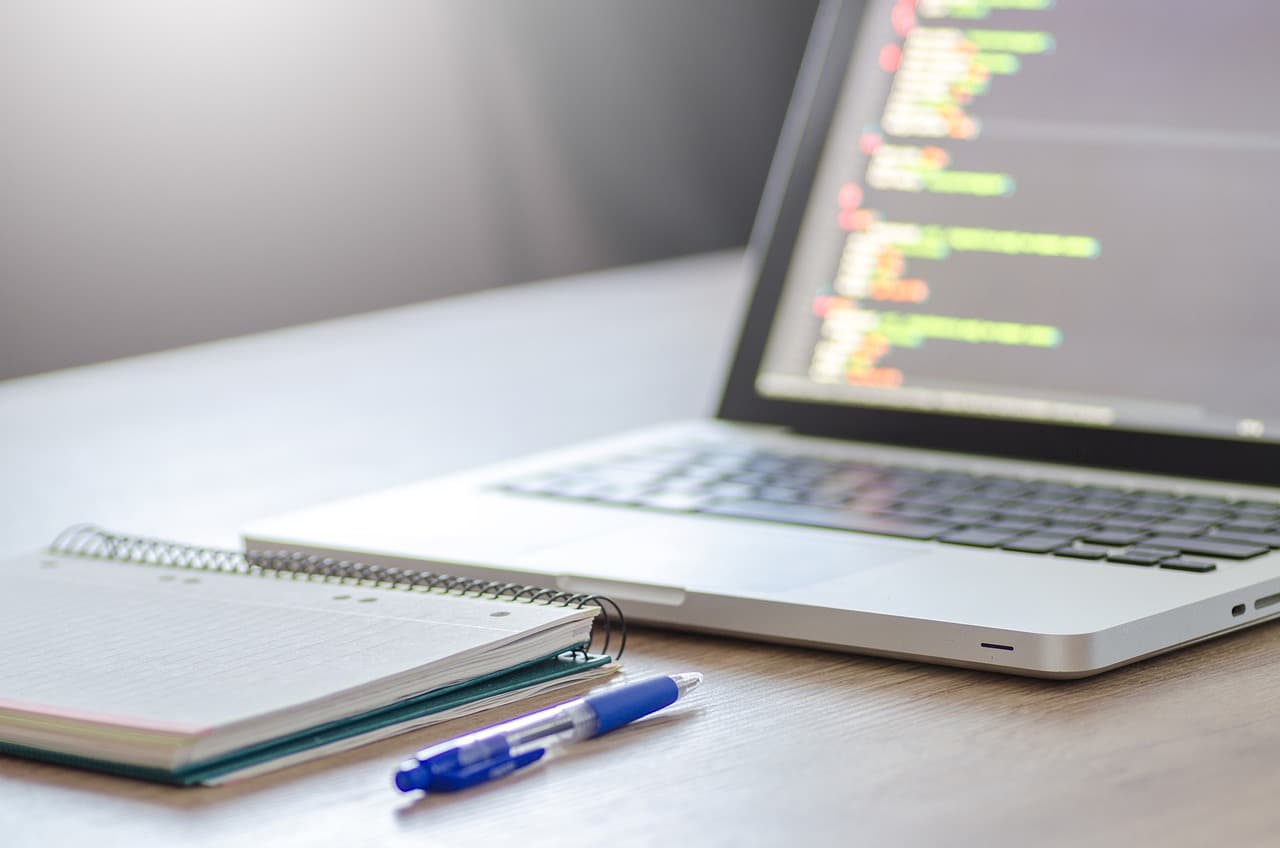 One blue pen resting next to a notebook with a blue cover and a Macbook computer behind them with multi-colored code displaying.