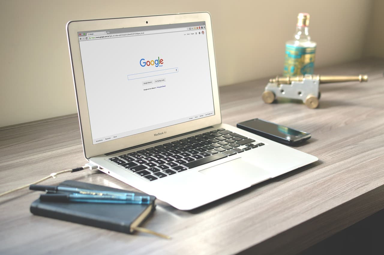 One silver Apple Macbook with the google screen on it next to a black notebook with two blue pens and an iPhone beside them all.