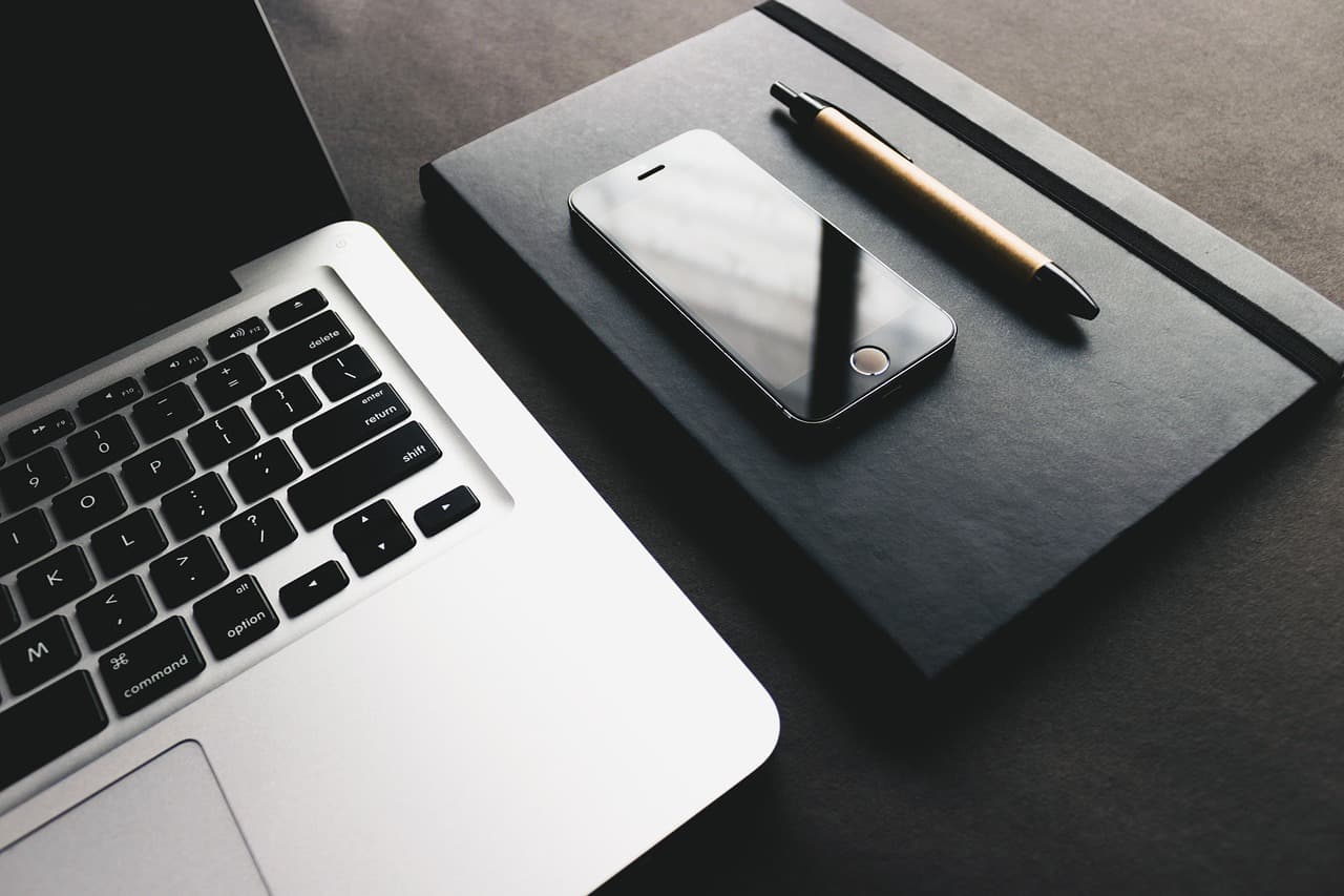 One black notebook with an iPhone that is turned off and a brown pen resting on top if it next to a silver macbook laptop.