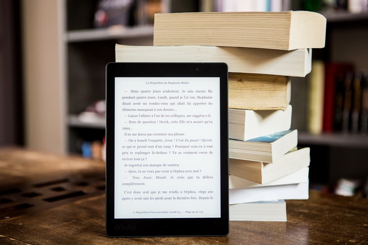 A digital tablet with a white screen with words on it in the back is leaning on a stack of nine books on a dark brown table.