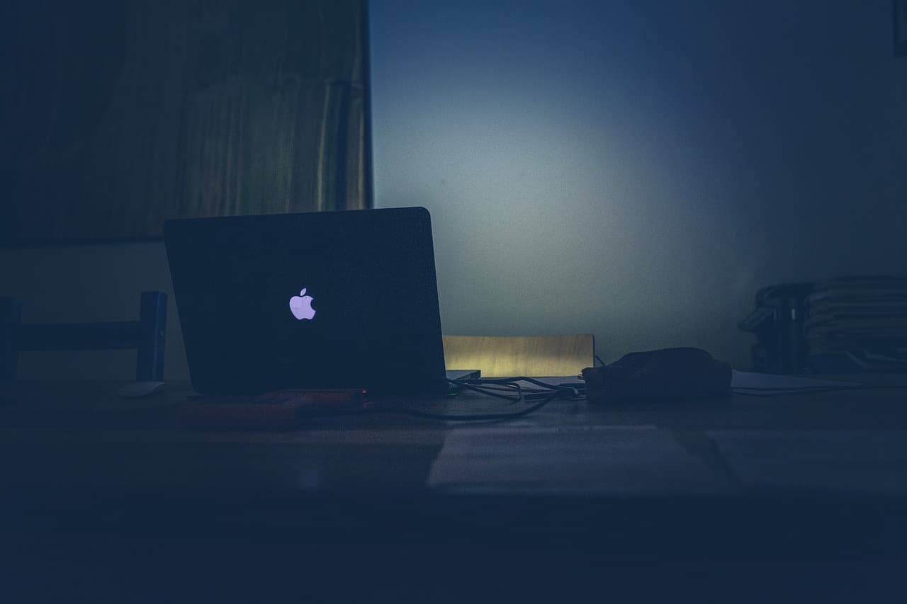 A silver Apple Macbook laptop lit up a dark room while being on a brown wooden table with chairs and black wires around it.