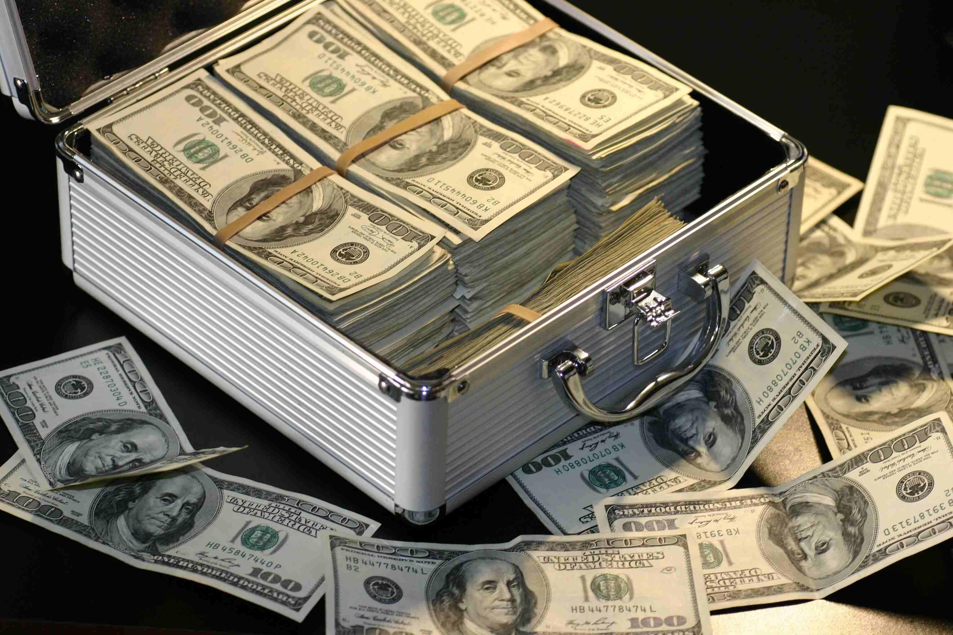 A briefcase full of one hundred dollar bill stacks with rubber bands on a black table with more bills surrounding the case.