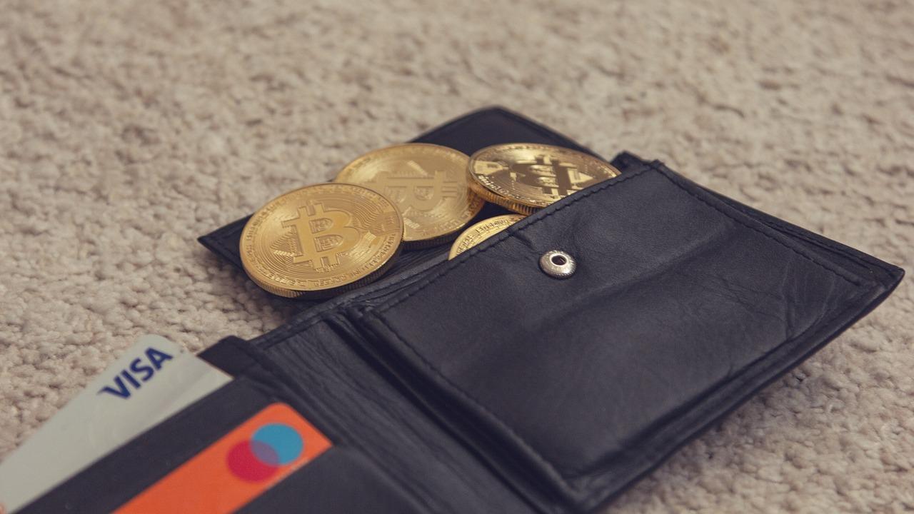 A darkly colored leather wallet with four golden coins coming out of it with the Bitcoin logos on them and two credit cards.