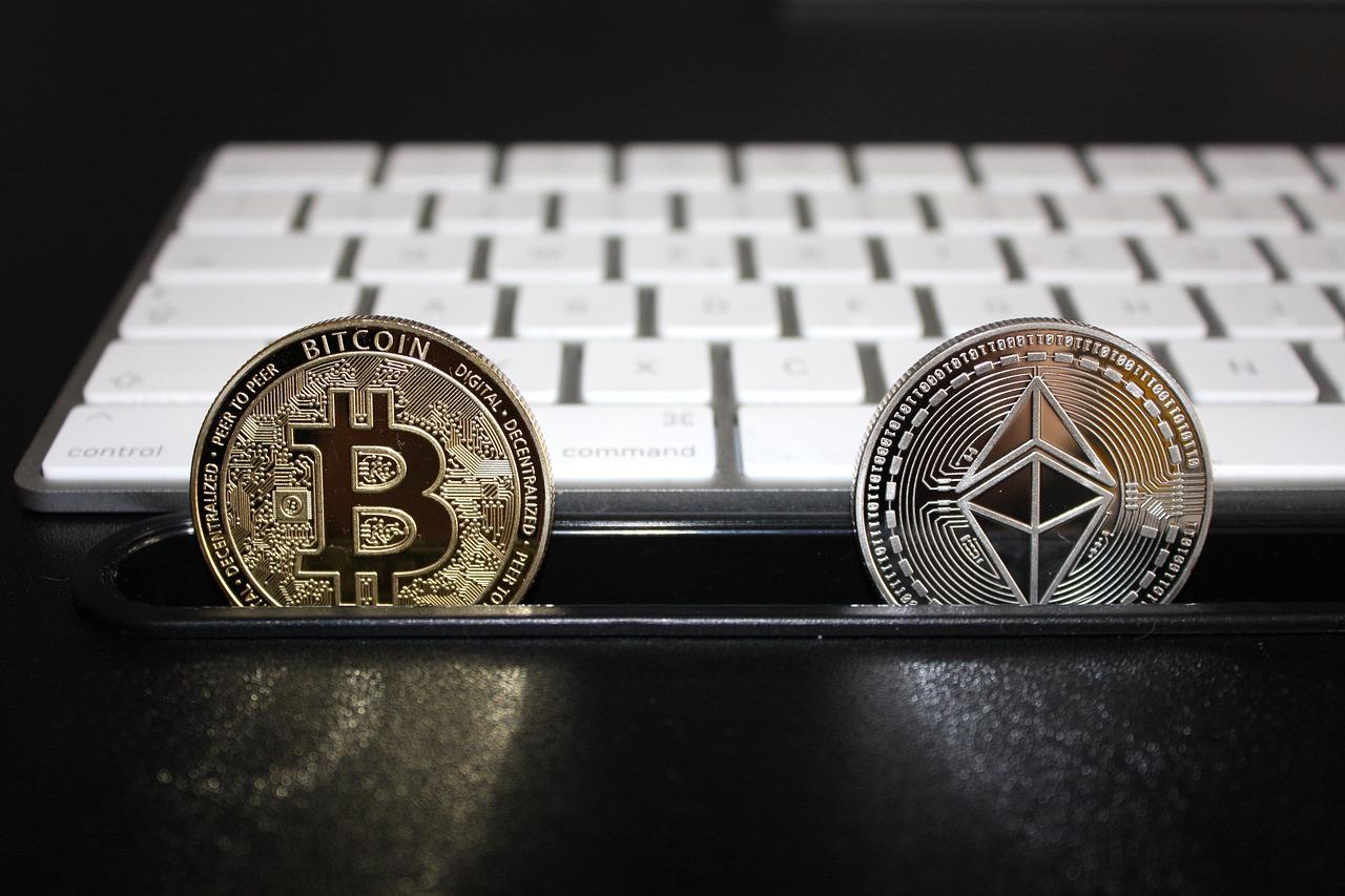 Two golden coins standing on their edge next to a white keyboard with the Bitcoin and Ethereum logos on them on a black surface.