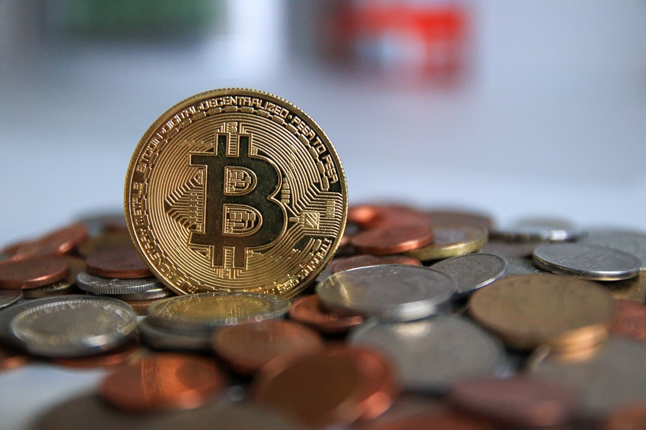 A golden coin with the Bitcoin logo on it sitting on top of a pile of other differently colored coins all on a white surface.