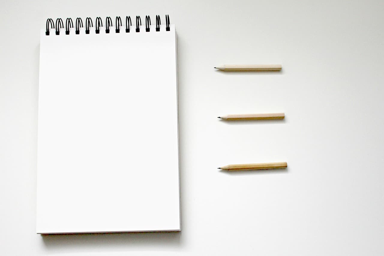 A white paper notebook with a black spiral holding it together on top on a lightly colored surface next to three pencils.