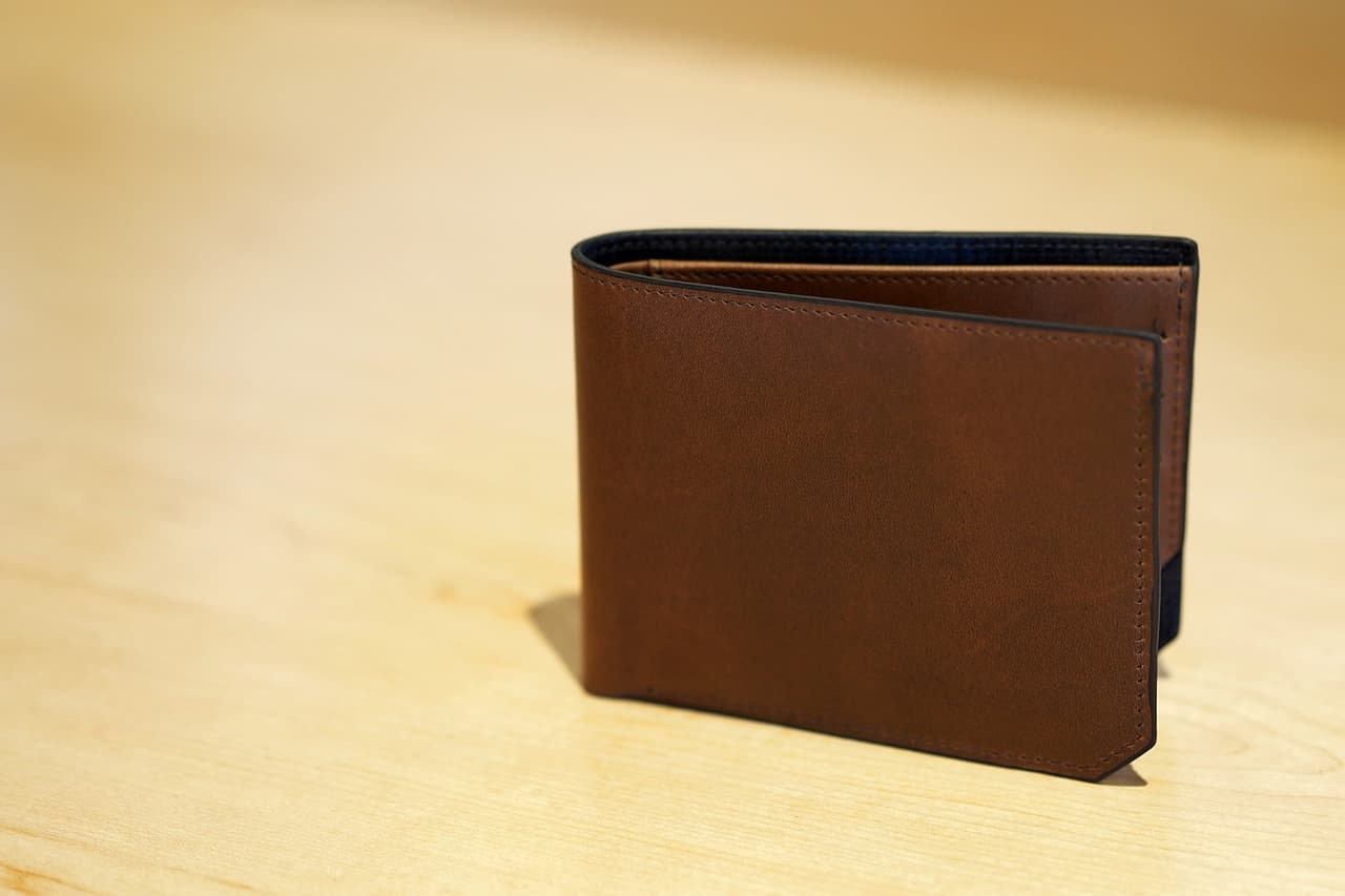 A dark brown wallet with nothing in it resting on it’s edge while slightly open on top of a lightly colored wooden table.