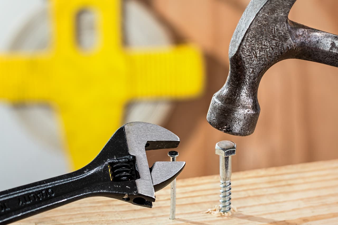 A silver hammer floating right above a bolt inside a piece of wood ready to smack it next to a nail with an adjustable wrench.