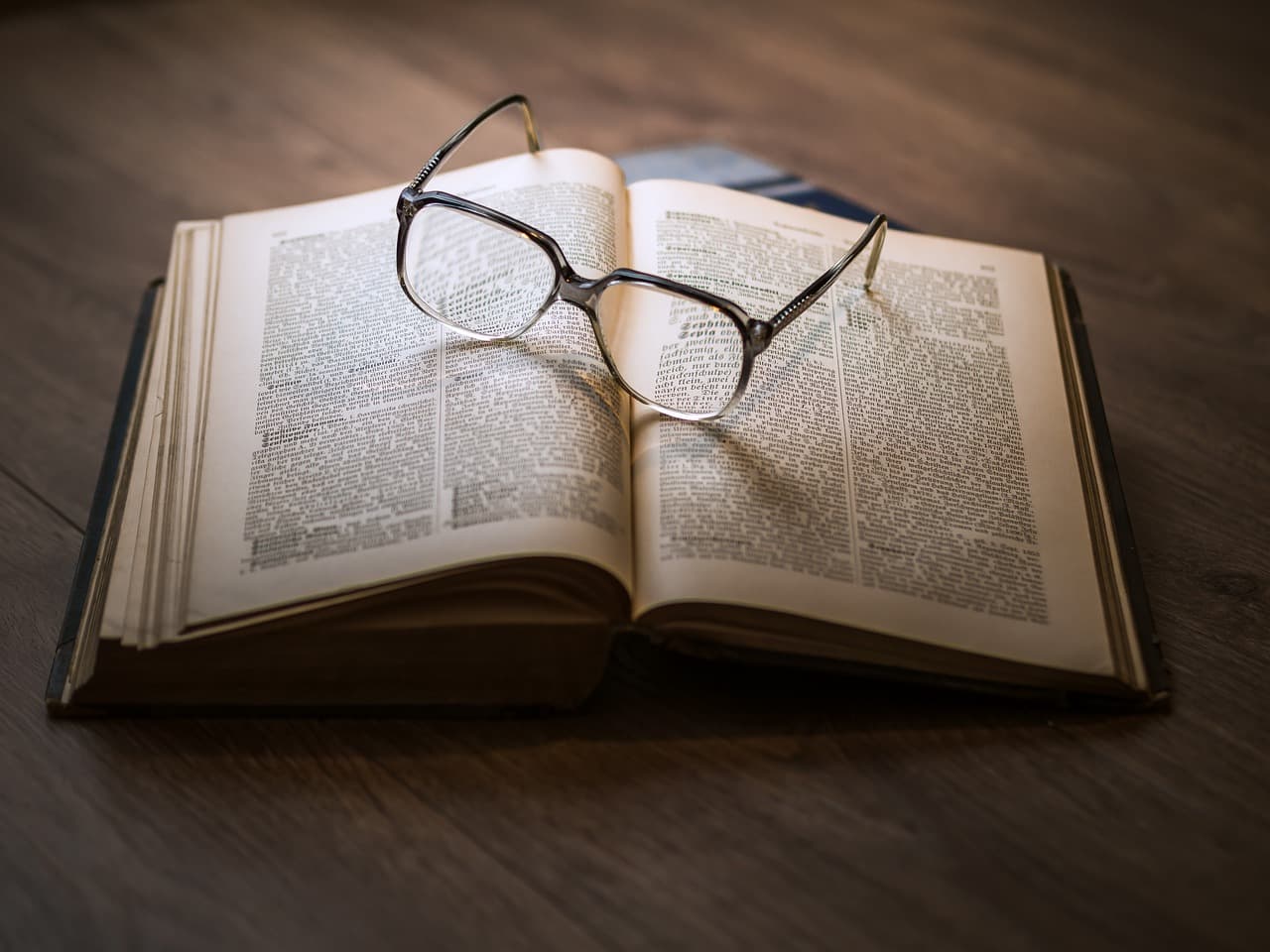 A book with a dark brown cover that is folded open with a pair of open glasses sitting on top resting on a dark wooden table.